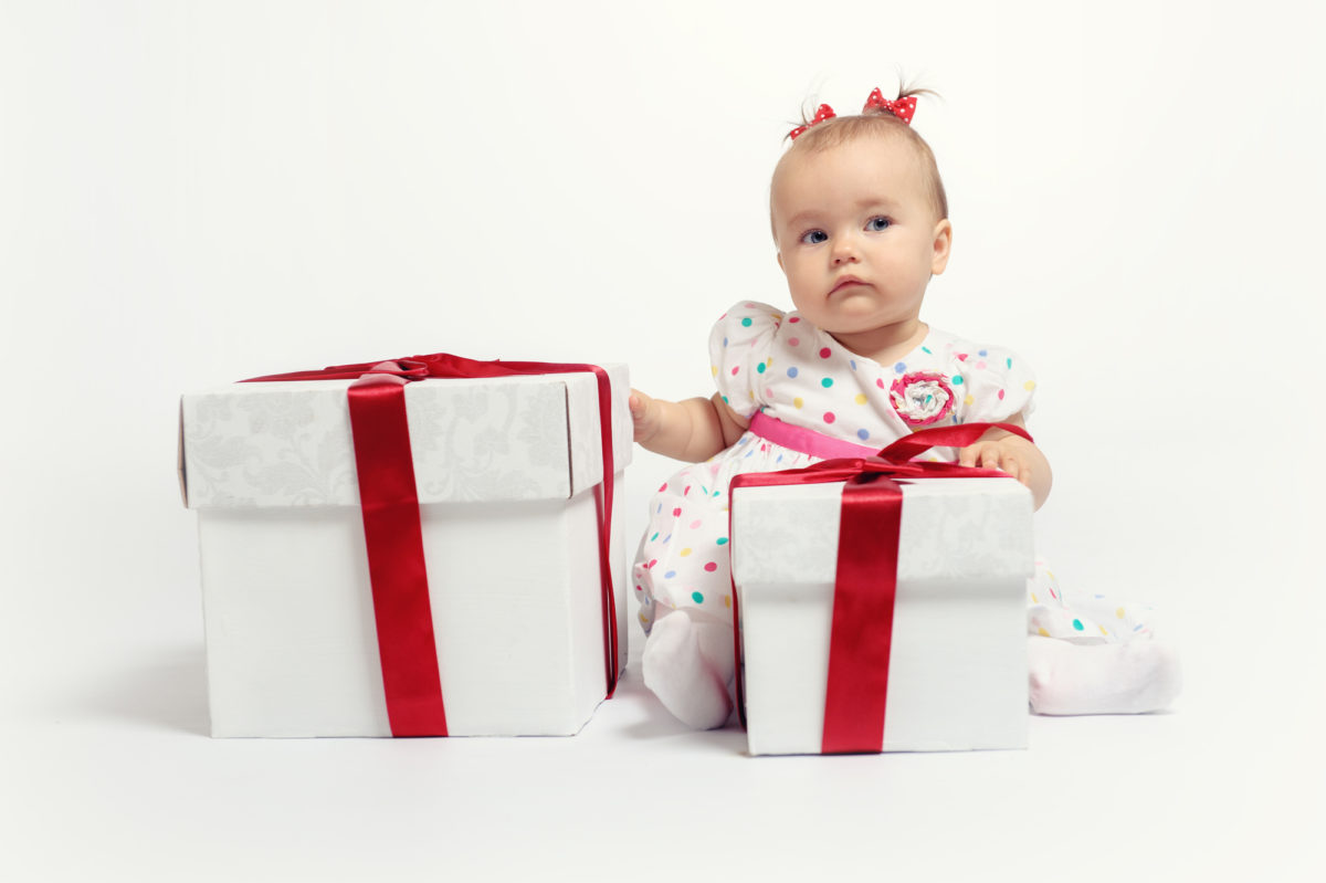 Kleinkind sitzt neben zwei großen Geschenken hinter einem weißen Hintergrund.